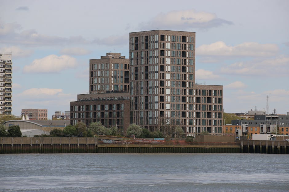 A modern, multi-story residential building with a stepped, tiered design made of dark brick and numerous large windows, situated along a waterfront with a calm river in the foreground. The surrounding