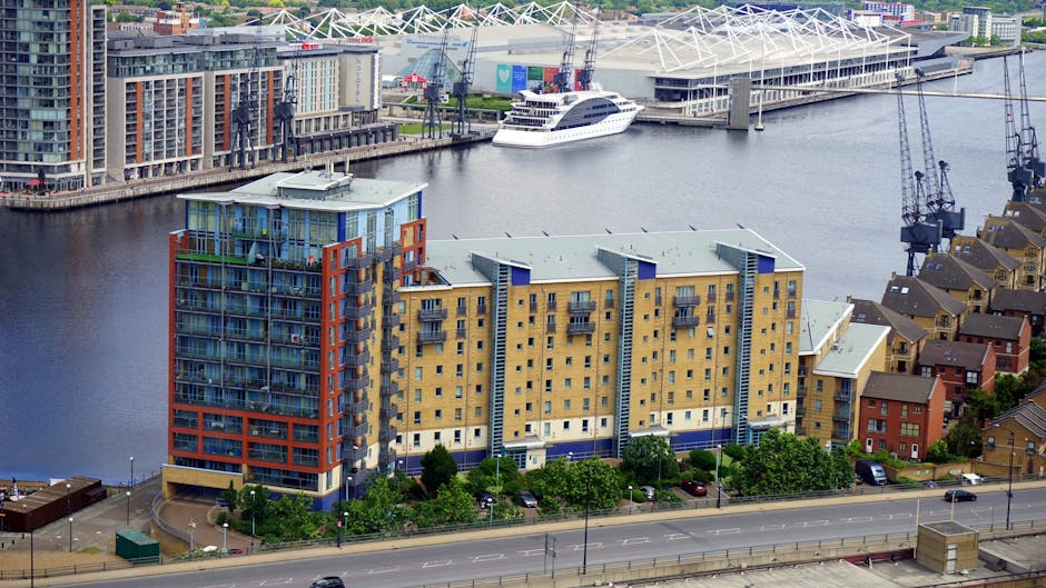 A modern, multi-story residential building with a stepped, tiered design made of dark brick and numerous large windows, situated along a waterfront with a calm river in the foreground. The surrounding